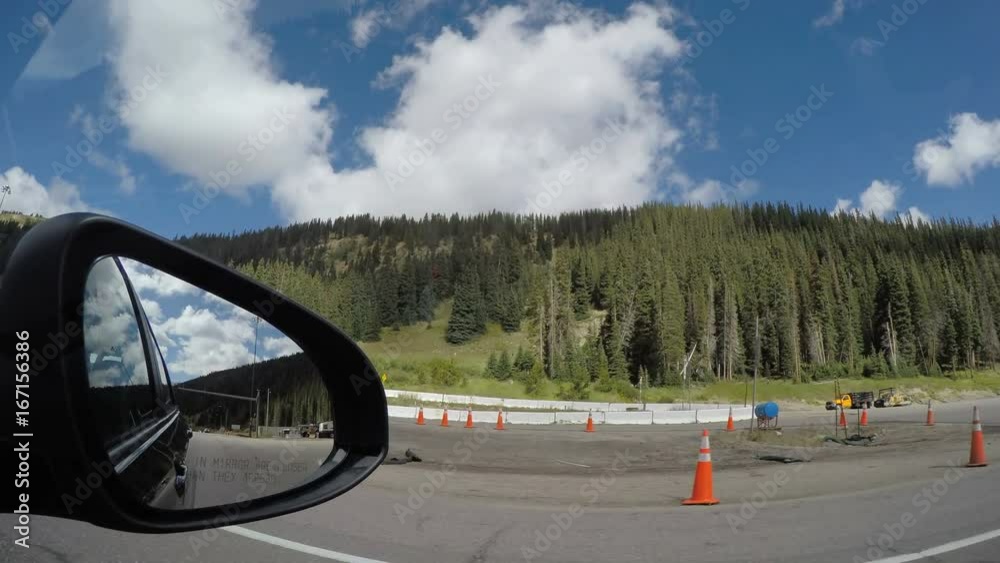 POV traveling driving moving by mountain area with cars and construction then entering an interior tunnel passageway double view through car side rearview mirror with blue sky clouds, pine trees and s