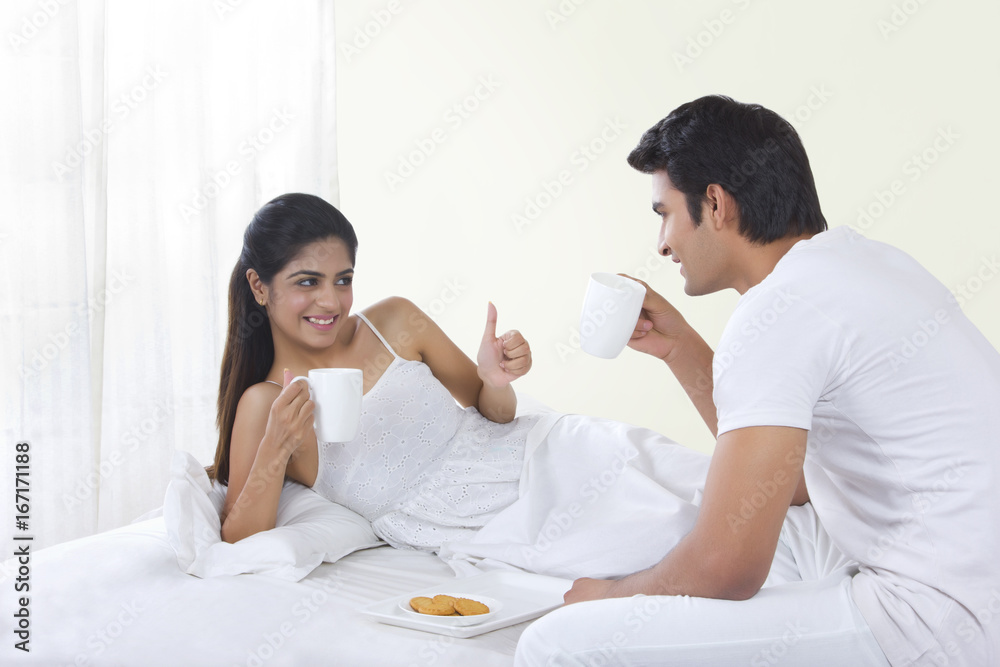 Happy young woman gesturing thumbs up while having coffee with man in bed Happy young woman gesturing thumbs up while having coffee with man in bed