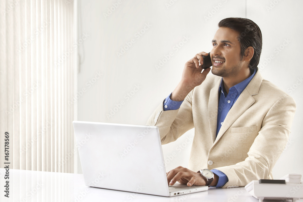 Young Indian businessman on call while using laptop in office Young Indian businessman on call while using laptop in office