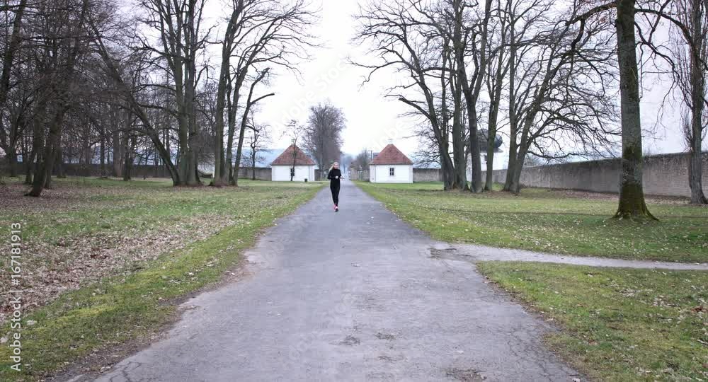 A woman taking a job in a park during autumn. Bare trees and fallen leaves cover the landscape beside. She runs towards the camera.