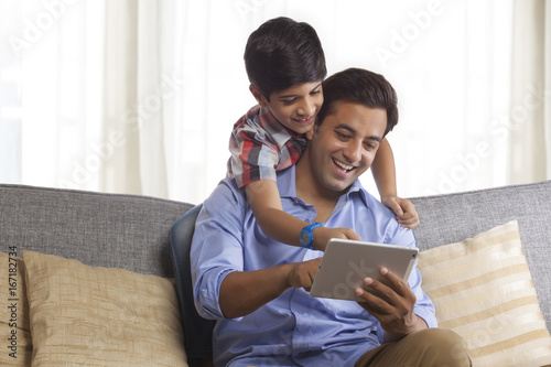 Smiling father using digital tablet sitting on sofa while his son looks from behind