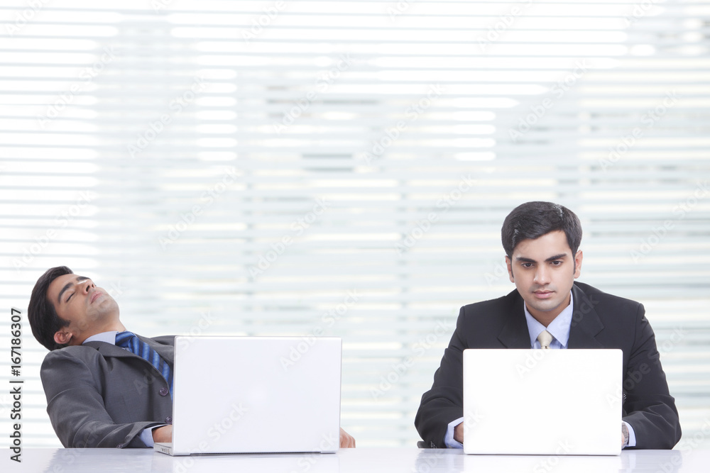 Young businessman working on laptop while colleague sleeping at desk in office Young businessman working on laptop while colleague sleeping at desk in office