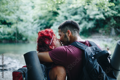 Young necking couple enjoying in hike. They walking through wild mountain river with a backpacks hugging and kissing. Rear view.