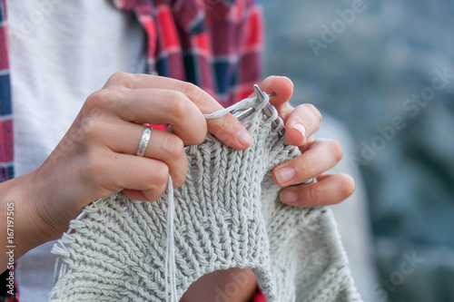 Close-up Young woman checkered red shirt and white t-shirt knits with knitting needles gray sweater from natural woolen threads, in the background stones