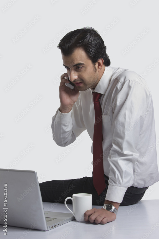 Young businessman answering mobile phone while looking at laptop over white background