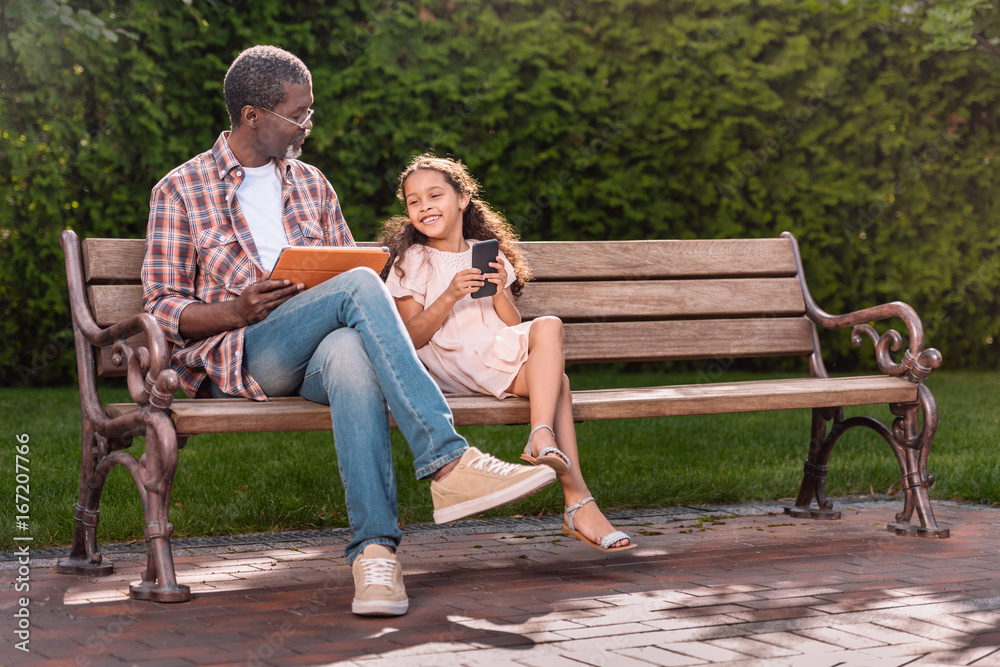 smiling african american girl and her grandfather using smartphone and digital tablet while sitting on bench in park smiling african american girl and her grandfather using smartphone and digital tablet while sitting on bench in park