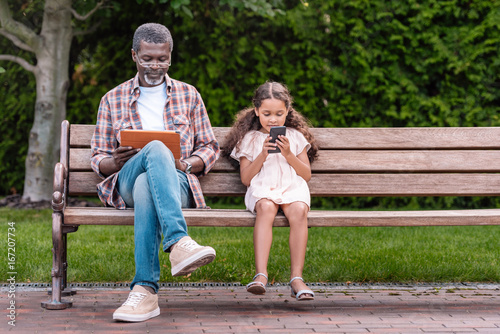 adorable african american girl and her grandfather using smartphone and digital tablet while sitting on bench in park