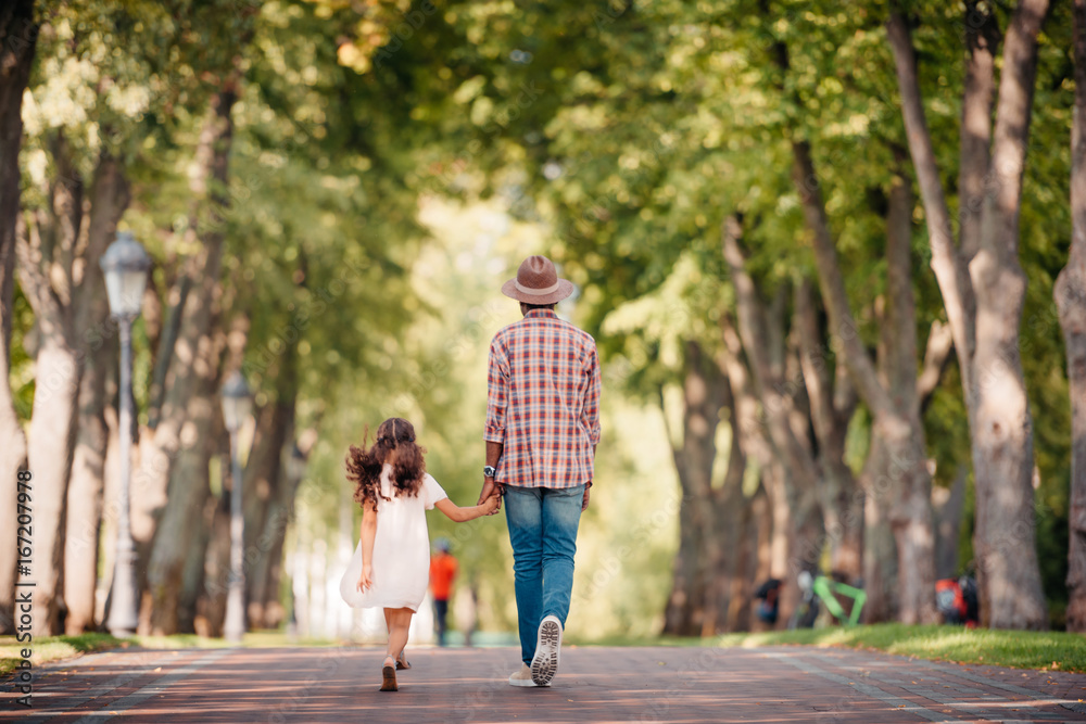 back view of african american girl holding hands with grandfather and walking in green alley