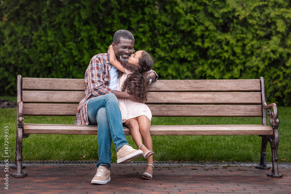 adorable african american girl hugging and kissing her grandfather while sitting on bench in park adorable african american girl hugging and kissing her grandfather while sitting on bench in park