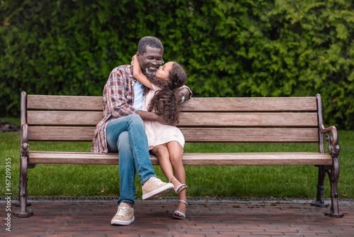 adorable african american girl hugging and kissing her grandfather while sitting on bench in park © LIGHTFIELD STUDIOS
