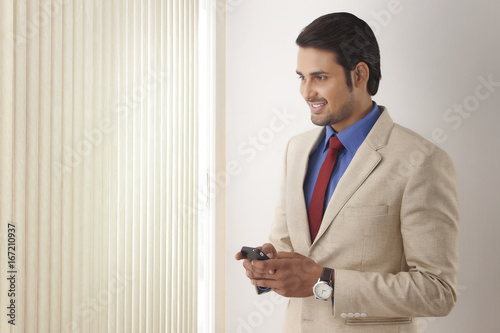 Young Indian businessman text messaging while looking through window blinds