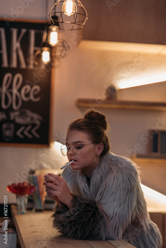 seductive fashionable young woman in fur coat smoking cigarette at bar counter