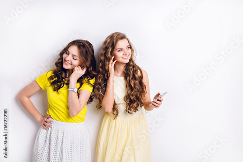 Studio photo of two standing smiling girls listening to music on a smartphone and having fun. Brunette wears white skirt and yellow T-shirt, and blond girl wears yellow dress.