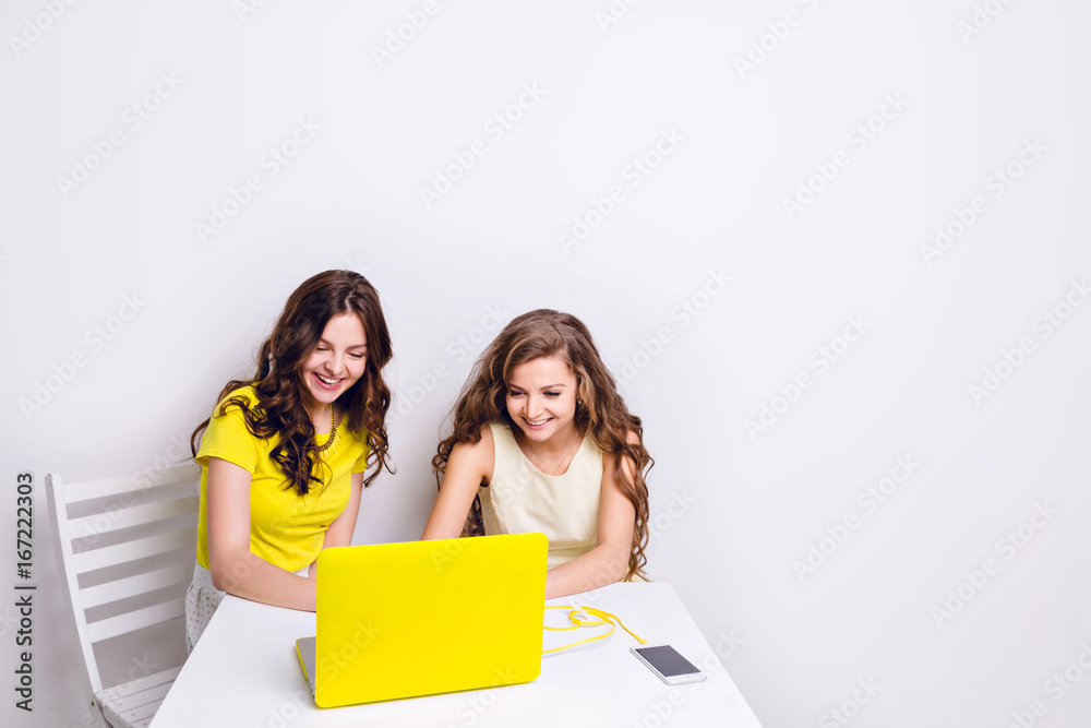 Two girls sit behind a laptop in yellow case. There is a smartphone on the table charging via yellow cable. Both girls are smiling and looking at the screen. One wears yellow t-shirt, another - dress. Two girls sit behind a laptop in yellow case. There is a smartphone on the table charging via yellow cable. Both girls are smiling and looking at the screen. One wears yellow t-shirt, another - dress.