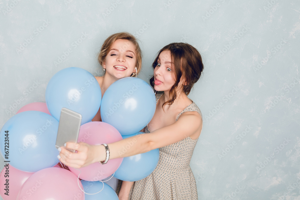Two girls having fun in a studio and playing with blue and pink baloons. Blond girl wears blue dress. Brunette girl holds smartphone listening to music on earphones. They take a selfie. Two girls having fun in a studio and playing with blue and pink baloons. Blond girl wears blue dress. Brunette girl holds smartphone listening to music on earphones. They take a selfie.