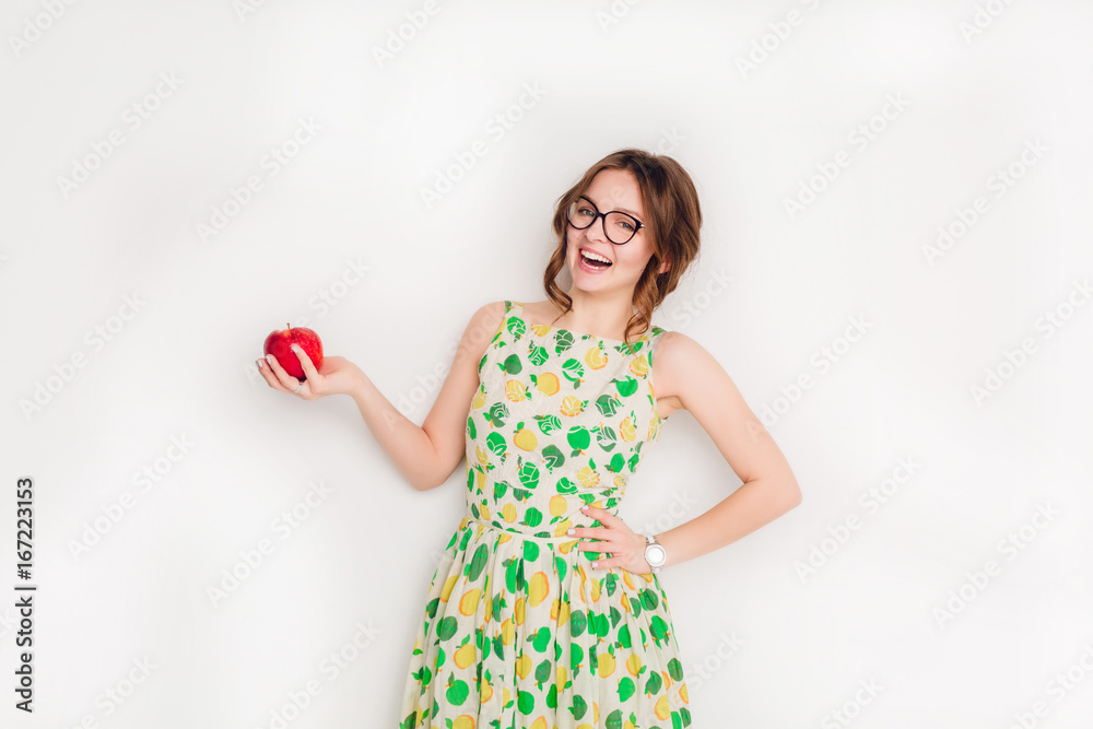 Studio shot of a smiling brunette girl smiling widely. Girl wears black glasses and yellow and green dress. She holds a red apple in her right hand. Studio shot of a smiling brunette girl smiling widely. Girl wears black glasses and yellow and green dress. She holds a red apple in her right hand.