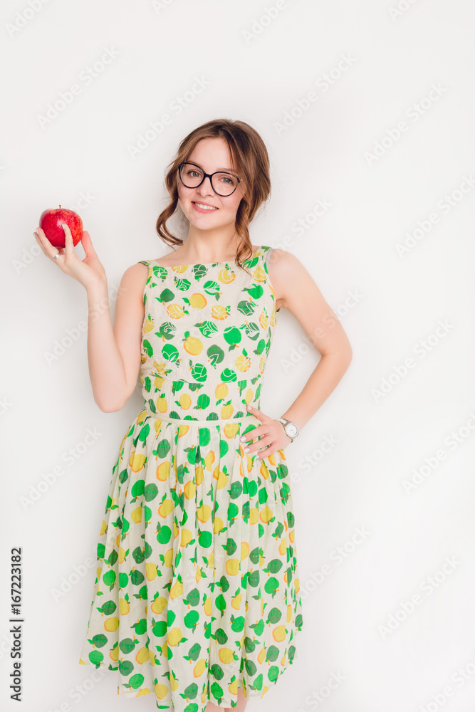 Vertical studio shot of a smiling brunette girl smiling widely. Girl wears black glasses and yellow and green dress. She holds a red apple in her right hand. Vertical studio shot of a smiling brunette girl smiling widely. Girl wears black glasses and yellow and green dress. She holds a red apple in her right hand.