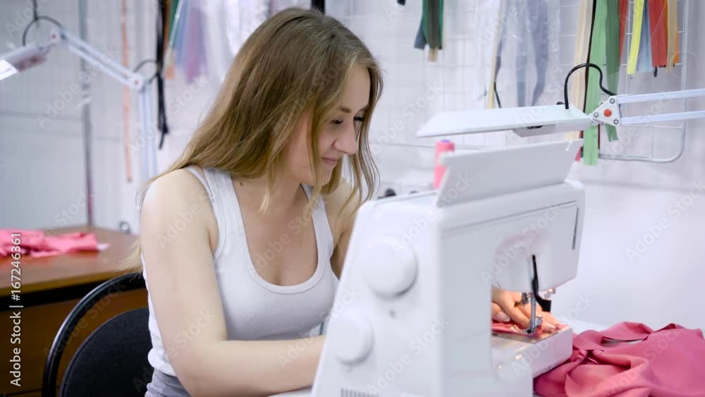 A young woman who works as a seamstress in a factory sews pieces of pink cloth on an electric sewing machine, in the future it will be a dress A young woman who works as a seamstress in a factory sews pieces of pink cloth on an electric sewing machine, in the future it will be a dress