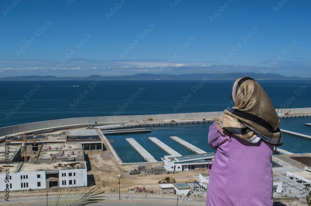 Fototapeta premium Nord Africa: una donna musulmana di spalle, di fronte al porto di Tangeri, guarda lo stretto di Gibilterra che unisce Oceano Atlantico e Mar Mediterraneo e separa il Marocco dalla Spagna