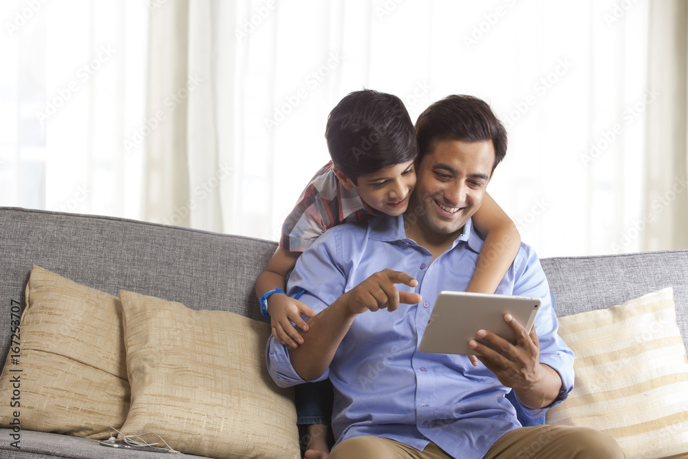 Smiling father using digital tablet sitting on sofa while his son looks from behind Smiling father using digital tablet sitting on sofa while his son looks from behind