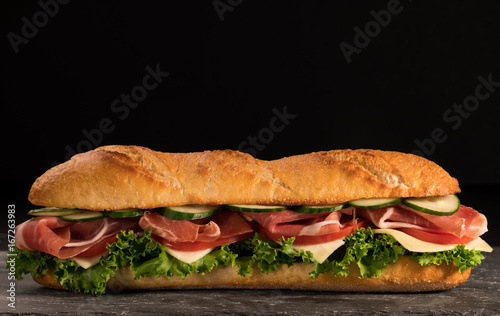 Huge crispy baguette deli sandwich with meat and vegetables. Close up. Black background.