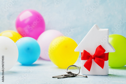 Small white wooden house decorated red bow ribbon with bunch of keys and balloons on light background. Housewarming, gift, moving, real estate or buying a new home concept. photo