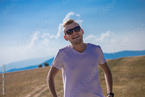 Handsome young caucasian blonde guy happy man with reflective black sunglasses laughing and smilingGrass field in the summer with mountains and blue sky and clouds in the background