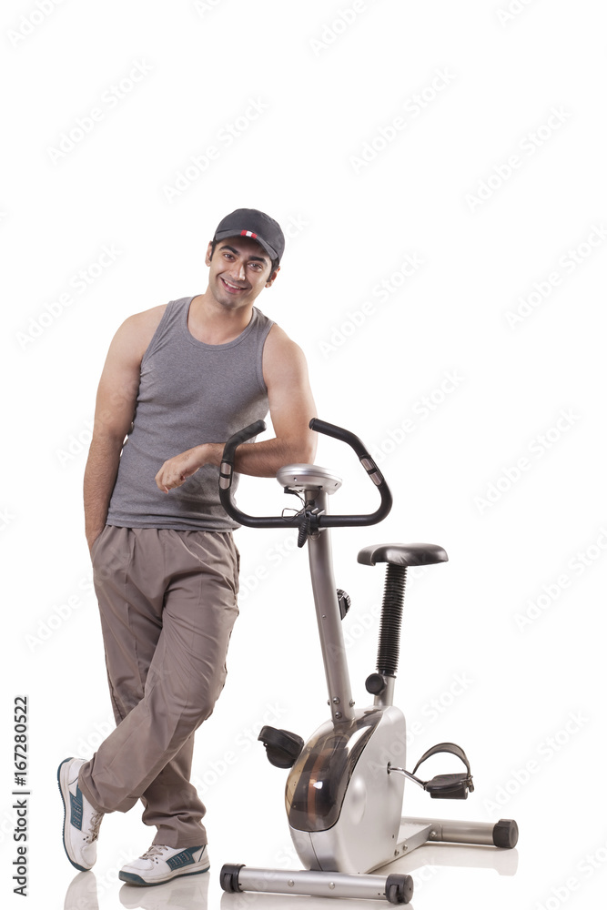 Full length of a young man standing next to an exercise bike over white background Full length of a young man standing next to an exercise bike over white background