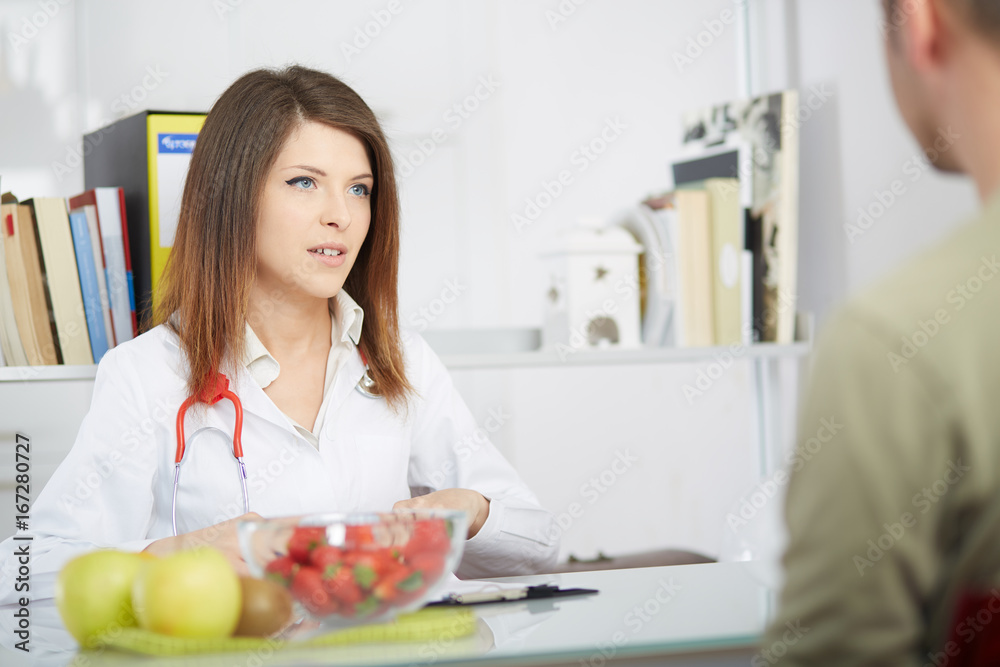 Smiling doctor nutritionist giving kiwi to male patient sitting at the desk with colorful fruits and vegetables Smiling doctor nutritionist giving kiwi to male patient sitting at the desk with colorful fruits and vegetables