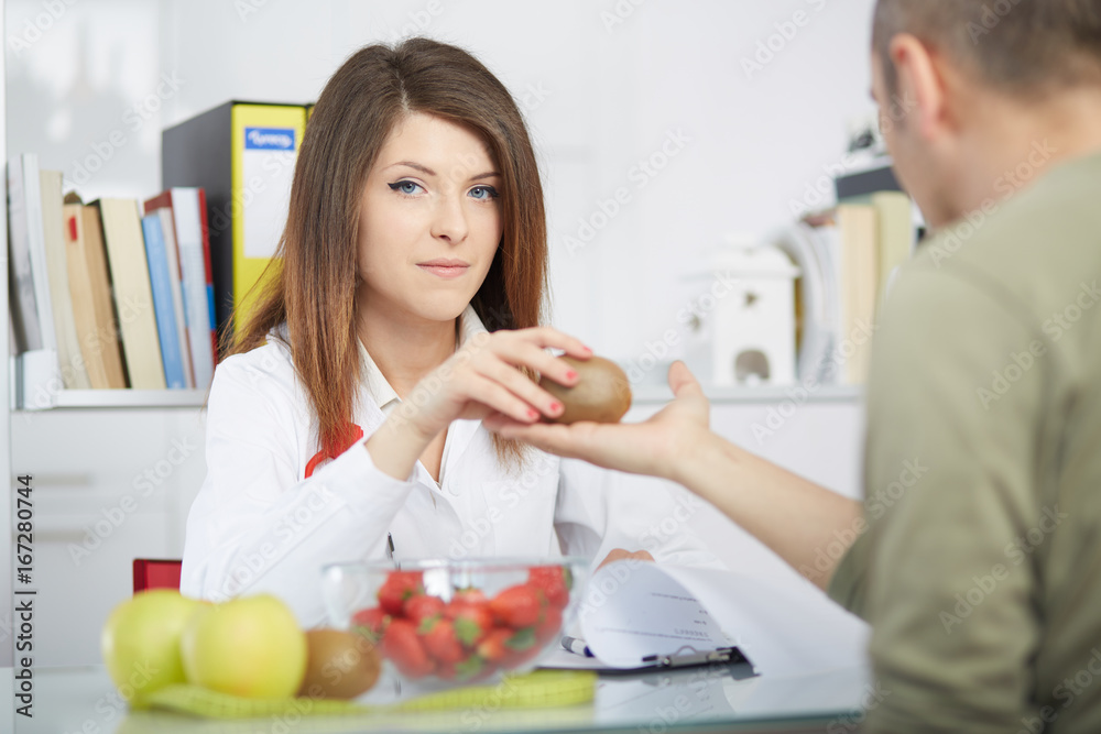 Smiling doctor nutritionist giving kiwi to male patient sitting at the desk with colorful fruits and vegetables Smiling doctor nutritionist giving kiwi to male patient sitting at the desk with colorful fruits and vegetables