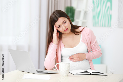 Beautiful young pregnant woman suffering from headache while working with laptop at home