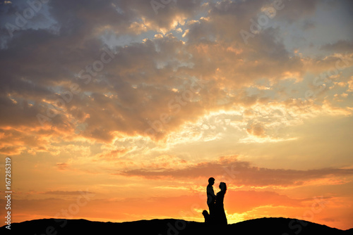 Silhouette of an unrecognizable loving couple with a dog standing on a sand in the background of the sunset.