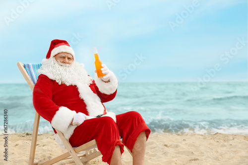 Authentic Santa Claus with bottle of drink relaxing in deck chair on beach