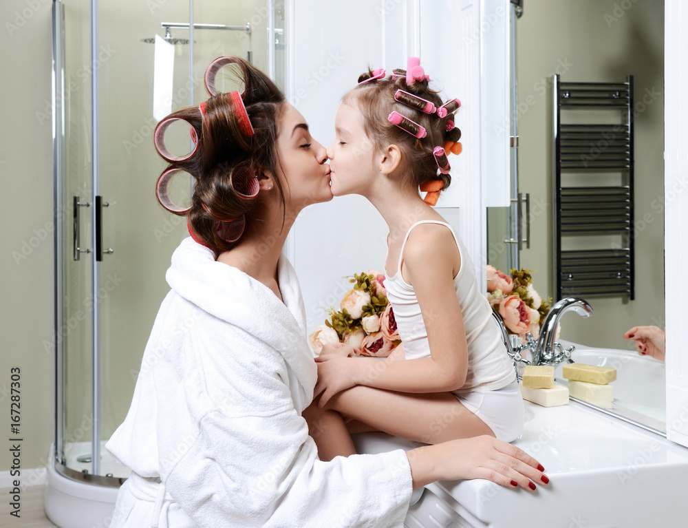 Young mother and daughter in curlers in a bath room happy smiling kissing g...