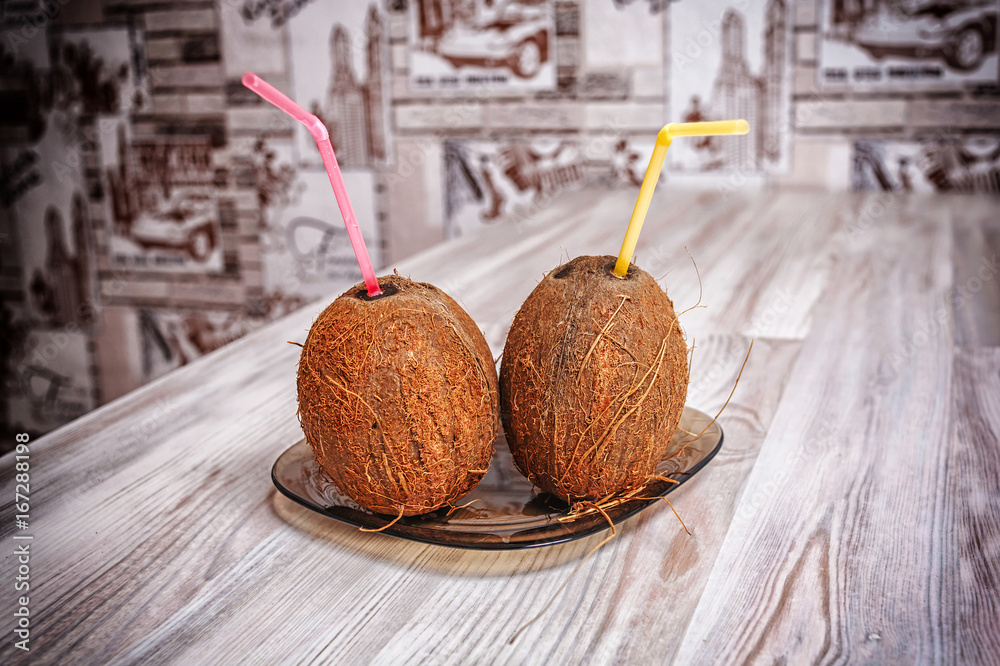 Two whole brown shell coconuts lie on a dark plate on a light table close up. In coconut nuts are inserted colored straws.