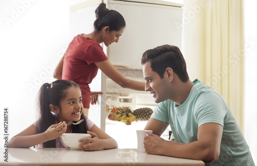 Father and daughter having breakfast while mother opening refrigerator