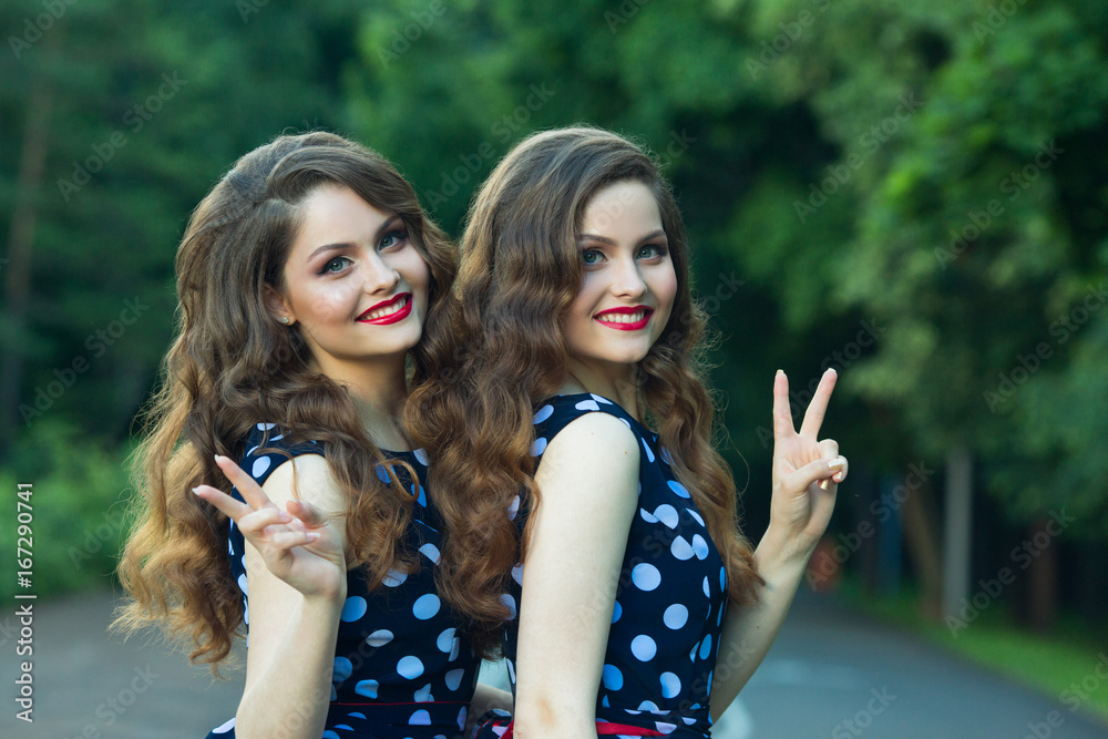 Two beautiful smiling girl sisters twins for a walk in summer park on a background of greenery with makeup and hairstyle Two beautiful smiling girl sisters twins for a walk in summer park on a background of greenery with makeup and hairstyle
