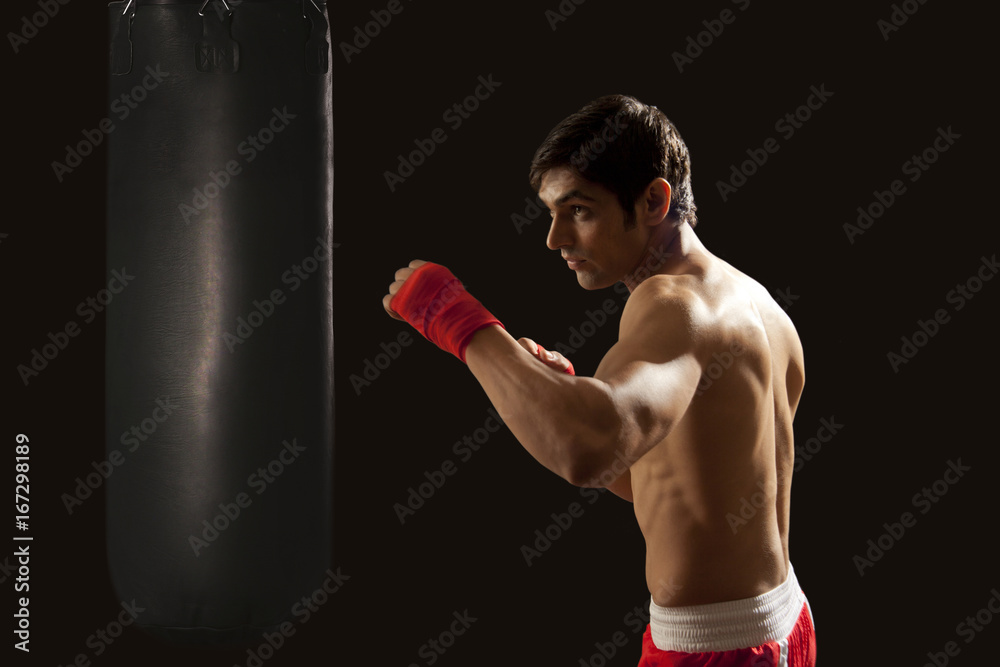 Side view of young man hitting punching bag isolated over black background