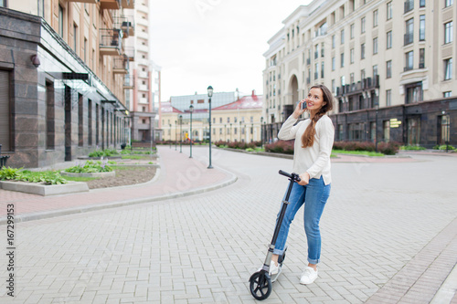 Beautiful young girl with long brown hair stopped while riding the scooter to talk to a friend on the phone on the background of the new residential quarter. Urban background