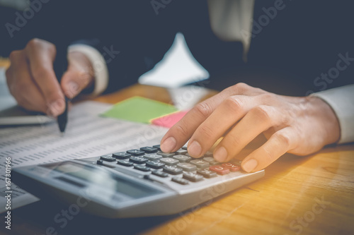 Close up business man hand holding pen and doing finance and calculate on wooden table about cost at home office. accountant calculator concept.