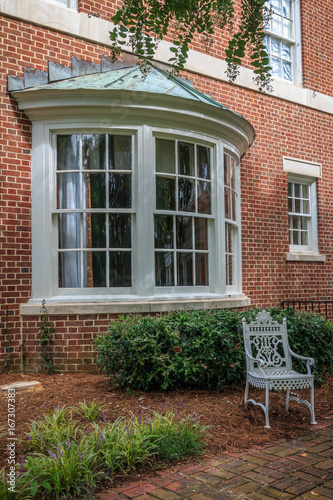 Curved exterior bay window with a metal roof is part of an southern mansion. A white classical iron chair sits in the yard in front of the bay window.
