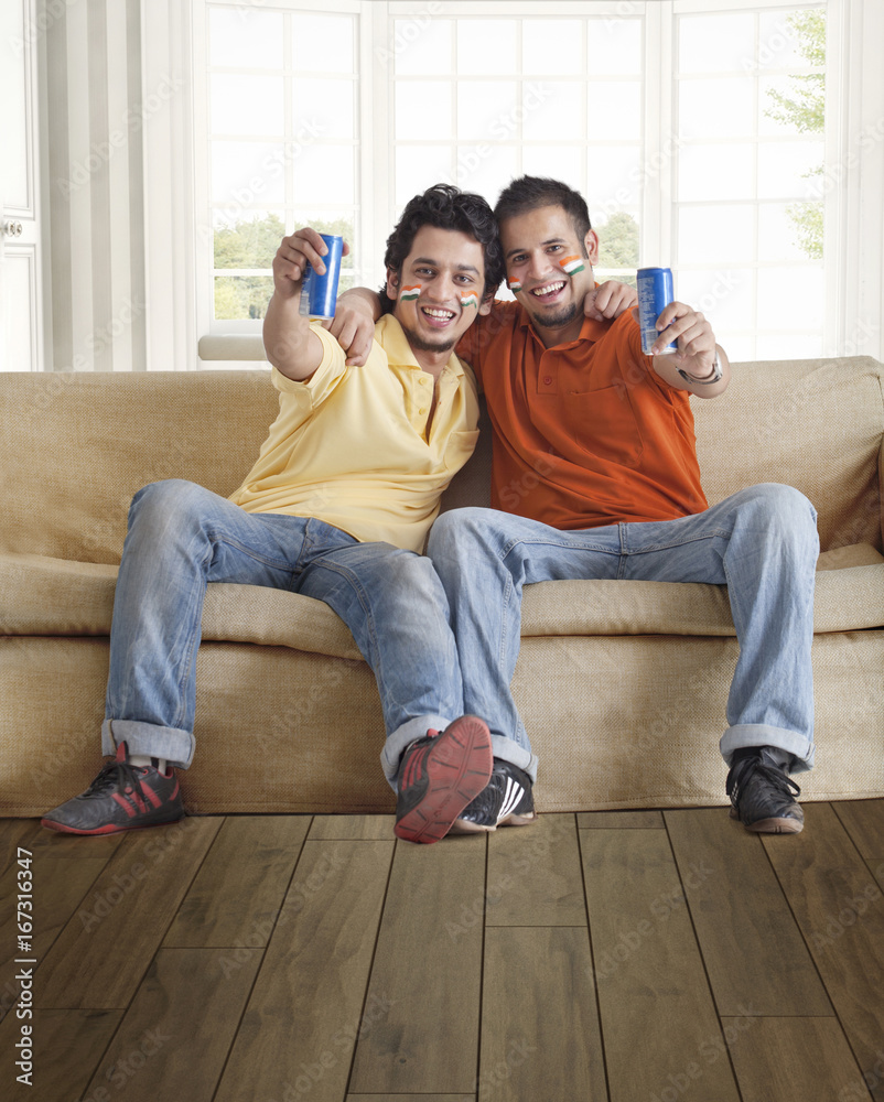 Portrait of happy young male friends holding tin cans while sitting comfortably on sofa at home 