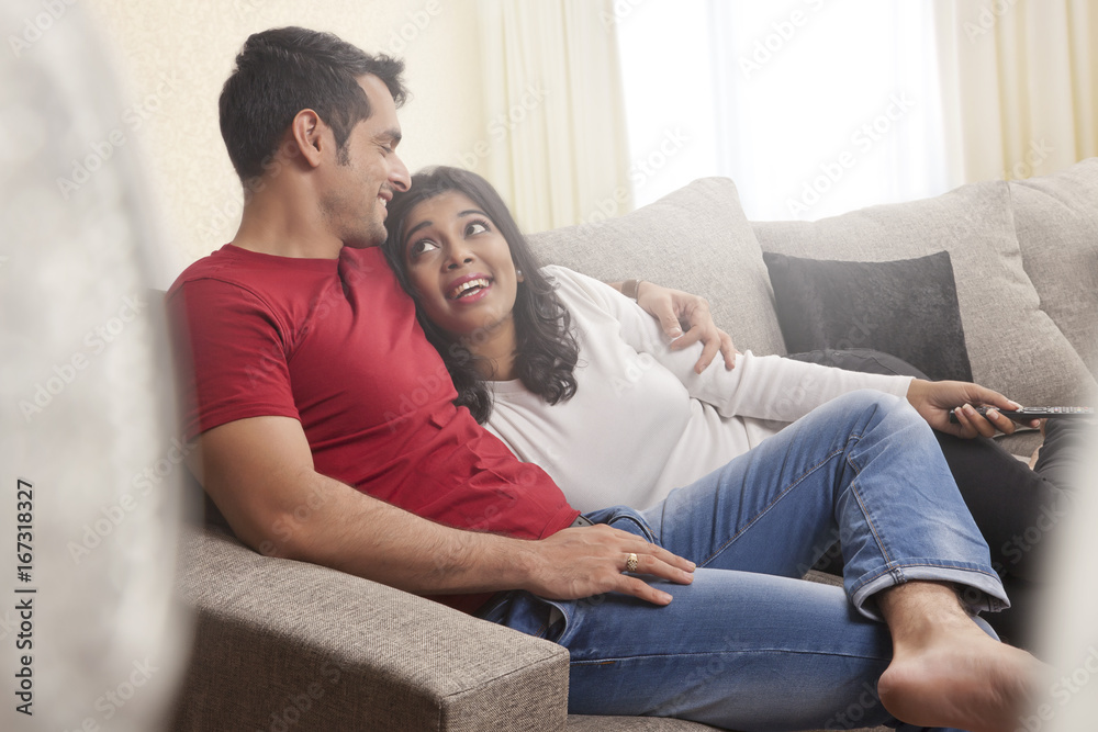 Smiling couple looking at each other sitting on sofa watching television Smiling couple looking at each other sitting on sofa watching television