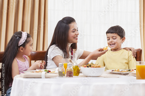 Cheerful mother feeding her son pizza with daughter sitting besides 