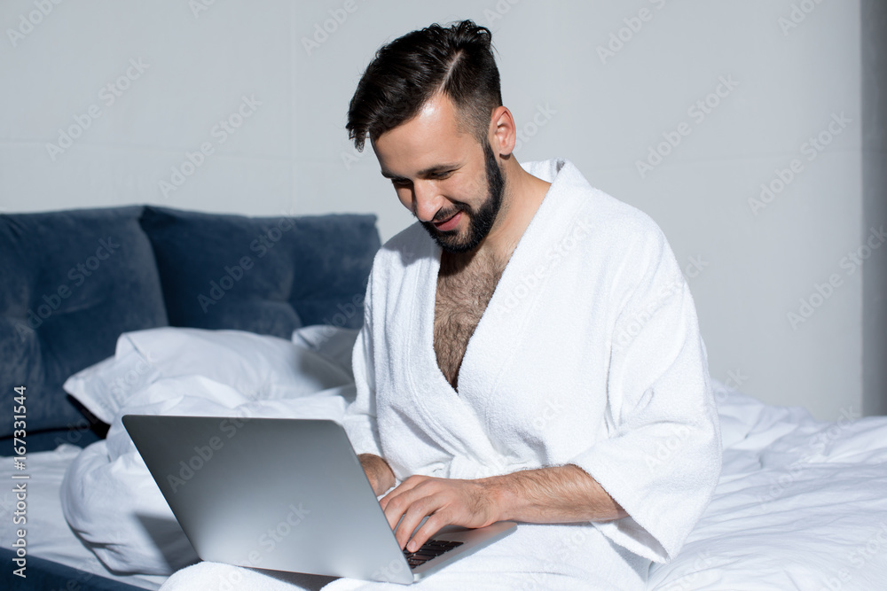 handsome young man in bathrobe using laptop while sitting on bed handsome young man in bathrobe using laptop while sitting on bed