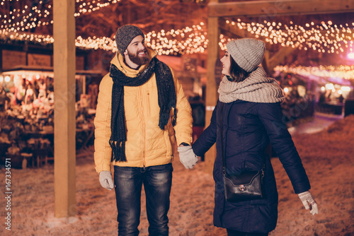 Happy carefree cute young married couple is walking in a christmas market, smiling, wearing warm winter outfits, many lights on the background