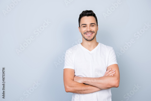 Success concept. Excited young stylish bearded brunet  guy in white t shirt standing on light background with crossed hands