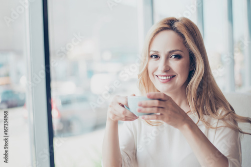 Good morning! Close up portrait of charming dreamy blond young lady drinking coffee. She is relaxed, in casual wear, in cafe, near the window