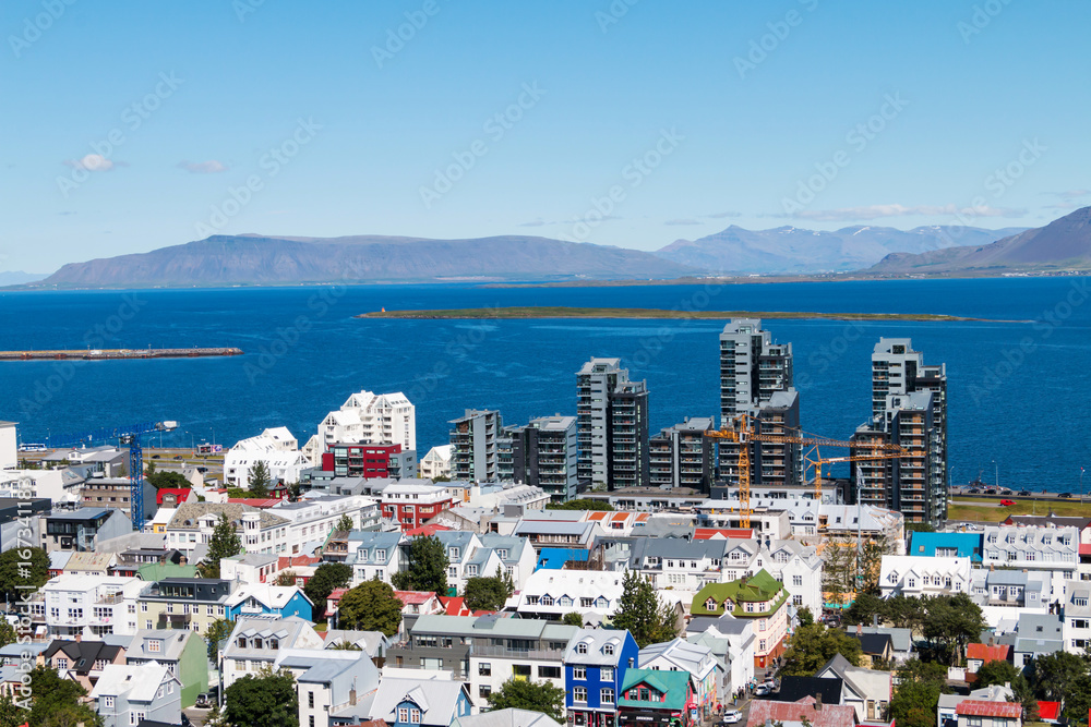 Aerial view on capital of Iceland - city of Reykjavik - ocean bay,  streets, houses, roofing, construction, cranes, mountains, horizon, blue skies, island - on sunny day.