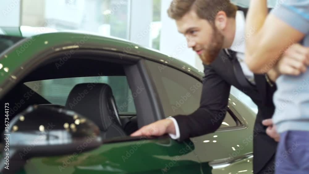 Young cheerful shop assistant showing new car to young casual man customer in showroom Young cheerful shop assistant showing new car to young casual man customer in showroom